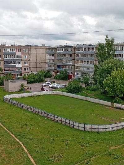 An aerial view captures a residential area in Rybinsk, Russia, featuring a multi-story apartment building and a small sports field. The building, constructed with concrete panels, displays rows of balconies and windows. In front of the building, a fenced-in sports field with a green grassy surface is visible, accompanied by a simple goalpost. A path winds through the grass. Several cars are parked in an adjacent lot. Surrounding the buildings and field are numerous trees and shrubs, indicating a green, possibly suburban, setting. The sky is overcast with a muted, gray tone.