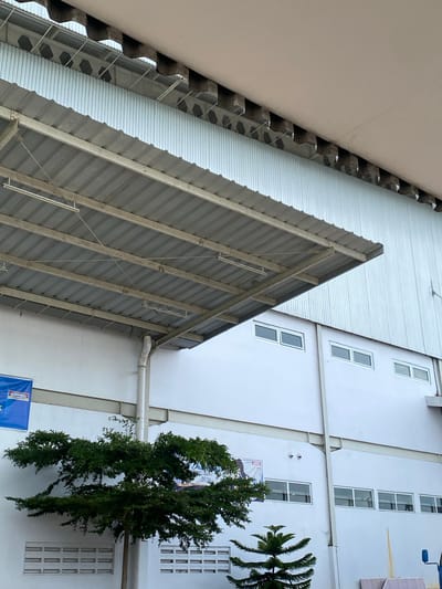 The image depicts the exterior of a white building, likely an industrial facility, in Gambut Barat, Indonesia. A corrugated metal overhang supported by beams extends from the building, casting a shadow on the wall below. A row of evenly spaced rectangular windows is visible on the building's facade. In the foreground, a lush green tree with dark foliage is situated to the left, partially obscuring some of the windows and a white rectangular vent on the wall. To the right of the tree, a smaller, spiky evergreen tree is visible. A blue banner with text and a logo is partially visible on the wall to the left of the tree. The sky is overcast and appears bright. The architectural style suggests a modern industrial or commercial structure.