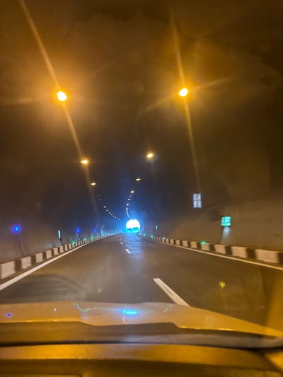 The image shows the interior of a road tunnel. The perspective is from the driver's seat of a vehicle, looking forward. The tunnel walls are dark, illuminated by a series of yellow lights mounted on the ceiling. The road surface appears wet, reflecting the lights. Along the sides of the road, there are black and white striped barriers and a line of small blue and green lights. In the distance, the tunnel opens up to daylight. There are no people or other vehicles visible in the immediate scene. Based on the provided metadata, the location is identified as the Tedotsminda tunnel in Georgia.