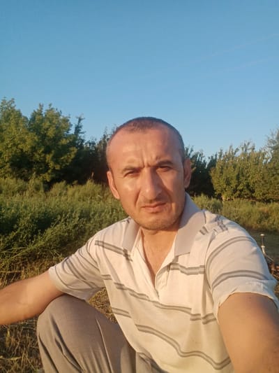 A man is captured in a selfie-style portrait outdoors. He has short, dark hair and is looking directly at the camera with a neutral expression. He is wearing a light-colored polo shirt with horizontal grey stripes and grey pants. The man is crouching down in a natural environment, with dense green foliage and trees in the background. The sky above is clear and blue, suggesting it is daytime, possibly late afternoon due to the warm lighting. The specific location, Romitan, Uzbekistan, places this scene within a rural or semi-rural setting.