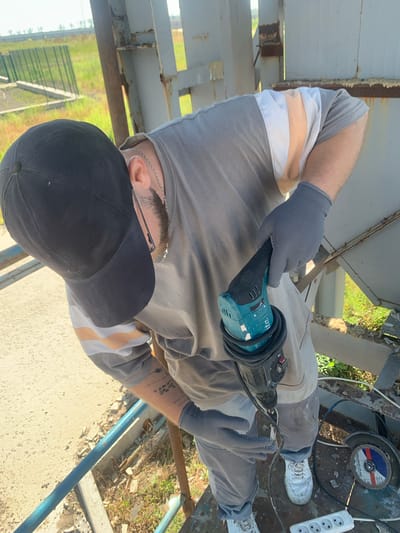 This image features a person in work clothes, wearing a black baseball cap and gray gloves, holding a blue and black power tool, possibly an angle grinder. They are positioned outdoors, with a background that suggests a construction or renovation site. Visible elements include metal structures, possibly scaffolding or beams, and some greenery. There is also a power strip with multiple outlets and a cutting disc. The person is wearing gray pants with light-colored stripes and white sneakers. The scene appears to be well-lit by daylight. The orientation of the image is rotated. The setting suggests an industrial or outdoor work environment in Pivdennoukrainsk, Ukraine.