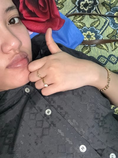 A close-up, slightly angled shot captures a person with fair skin. The person's face is visible from the forehead to the mouth, with one eye open and looking towards the camera. Their hand is positioned near their face, adorned with a gold bracelet and two gold rings on the ring and pinky fingers. They wear a black button-down shirt. The background consists of a floral-patterned surface with blue and red accents.
