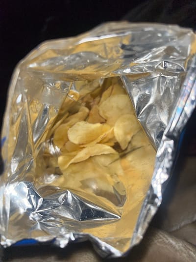 The image displays an opened snack bag, identified as containing potato chips, located in Nuseirat, Palestinian Territory. The bag's interior is visible, characterized by a crinkled, highly reflective silver-foil lining. Within the bag, numerous light golden-brown, irregularly shaped potato chips are present. At the bottom of the bag, a significant accumulation of yellowish-orange powder, consistent with seasoning or potato residue, is observable in the creases. The upper edges of the bag are open and somewhat distorted from its original sealed state. The background outside the bag is dark and out of focus, providing no further contextual details. No individuals or specific actions are depicted.