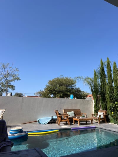 The image displays a residential backyard scene in Kfar Yona, Israel, under a clear blue sky, indicating daylight. A rectangular swimming pool with dark-colored tiles occupies the lower and central portion of the frame, containing clear water. Floating within the pool are a yellow and blue pool noodle, a purple pool noodle, and a blue and white inflatable float positioned near the left edge. A striped towel, possibly blue and white, is partially visible draped over an object in the immediate foreground to the left.
Behind the pool, a light gray concrete wall spans the width of the yard. Adjacent to the pool on the right, a patio area features a set of wooden outdoor furniture, comprising a two-seater bench with a light blue cushion, a rectangular coffee table, and a single armchair with a light blue cushion. To the left of the furniture set, a light blue tarp covers an indistinct object on the ground, with a long brown pole resting nearby.
Above the concrete wall, various trees are visible. To the left, a deciduous tree with sparse green foliage stands, with hints of distant buildings and a black and white sign appearing above the wall. Centrally, a large, dense green tree is prominent. To the right, several tall, slender green conifer trees (possibly cypress) and a palm tree are visible, along with a small blue circular sign depicting a white figure, positioned above the wall. No individuals are present in the scene.