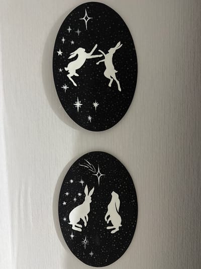 The scene, located in Vniissok, Russia, displays two oval-shaped art panels mounted vertically against a light-colored, vertically textured wall. Each panel features white silhouettes of hares and stars on a dark, speckled background.
The upper panel shows two white hares in dynamic poses against the dark, stippled expanse. The hare on the left is depicted mid-leap, facing right with its front paws extended. The hare on the right is also airborne, reaching its left foreleg towards the left hare's outstretched paw, suggesting an interactive dance. Several white, stylized stars, including one prominent four-pointed star, are scattered across the dark background.
The lower panel features two white hares in contemplative or observing poses. The hare on the left is seated on its haunches, facing right and gazing slightly upwards. The hare on the right stands on its hind legs, looking directly towards the top of the panel. The dark, stippled background includes multiple white, stylized stars. A central star at the top of this panel features a radiating tail, suggesting a shooting star.
The wall exhibits a consistent vertical striation texture, indicative of wallpaper or a similar finish, and appears off-white or light grey. Both art panels cast faint, diffused shadows on the wall, indicating they are slightly raised from the surface. The lighting is even, illuminating the wall's texture and the details of the artwork.