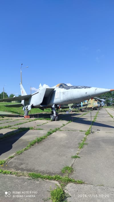 The image shows a static display of military aircraft outdoors on a sunny day. In the foreground, a light grey fighter jet with the number "07" on its side is parked on a concrete tarmac. The jet has wings, a visible cockpit, and landing gear. Red and orange cones are placed near the landing gear, and thin ropes are strung between posts along the tarmac. Behind the fighter jet, other aircraft, including a helicopter with a green and tan camouflage pattern, are visible further down the tarmac. The background consists of a grassy area with trees and a bright blue sky. The environment appears to be a museum or an outdoor exhibition area for vintage aircraft. The metadata at the bottom of the image indicates "REDMI NOTE 9 PRO AI QUAD CAMERA" and a date of "2026/7/15 10:20".