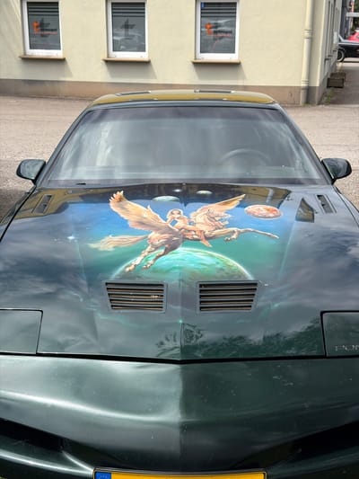 The image captures the front of a dark green Pontiac Firebird adorned with a custom airbrushed mural on its hood. The mural depicts a mythological scene set in space, featuring a winged horse with a rider, along with celestial bodies like planets and stars, painted against a gradient background of blues and greens. The car is parked outdoors, in front of a pale yellow building with two windows on the upper facade, each featuring a window decal with an orange logo. To the right of the building, a dark-colored car is partially visible. The foreground of the image shows the car's headlights, hood vents, and the bottom edge of the windshield, with a yellow license plate visible at the very bottom. The scene is located in Reckange-sur-Mess, Luxembourg, as indicated by the license plate. The surrounding environment appears to be a street or parking area, with a pavement in the foreground and a building with windows and architectural details like a downspout in the background.