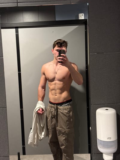 An athletic adult male is depicted in a mirror selfie within an indoor setting, consistent with a public restroom or gym changing room. The specific location within Chantepie, France, is not identifiable from the visual evidence. The male, appearing to be in his 20s or 30s, stands shirtless, displaying a muscular physique with visible abdominal definition. He has short, dark, curly hair and a light mustache, and wears a thin metallic chain with a small crucifix pendant. He holds a black smartphone in his raised left hand, angled to photograph his reflection. His right hand clutches a crumpled light-colored t-shirt. He is dressed in olive or dark brown cargo pants, with patterned boxer briefs visible above the waistband. The background reflected in the mirror features a light grey partition wall with a black vertical frame to the left, and a dark grey tiled wall to the right. A white, wall-mounted Tork brand dispenser is affixed to the dark tiled wall. A reflection of another white smartphone is visible in the upper right corner of the mirror.