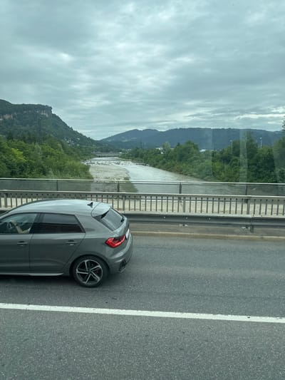 The image shows a grey hatchback car driving on a road next to a river and a bridge. The car is in the foreground, partially obscuring the view of the bridge and river. The river is wide and appears to be shallow with a rocky bed. The bridge is made of metal and concrete and spans the river. In the background, there are lush green trees and hills, with mountains in the distance. The sky is overcast with grey clouds. The scene appears to be a scenic location, possibly in a rural or mountainous area. While Bregenz, Austria is mentioned, there are no distinct landmarks or contextual clues in the image that definitively place it in that specific location.