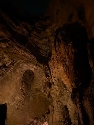 The image shows the interior of a cave with rough, textured rock formations illuminated by artificial lighting. The rock walls display layers and striations, with areas of deep shadow contrasting with patches of warm orange and brown light. In the lower left corner, a dark, rectangular object, possibly an entrance or a piece of equipment, is partially visible. The overall environment is dark, cavernous, and appears to be a natural geological formation. There are no people or animals visible in the image. The image does not provide specific visual cues to confirm its location in Guttapadu, India.