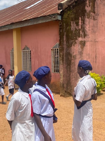 Three young individuals are pictured, all wearing similar uniforms consisting of white robes, navy blue hats, and colored sashes or neckties. They appear to be in a courtyard or outdoor area, perhaps in a location in Turu, Nigeria, given the nature of the request. Behind them is a building with pinkish-red walls and what seems to be a rusty corrugated metal roof. The building has small, grated windows. The ground is a sandy or dirt surface. The children seem to be standing in a line, and the light suggests it is likely daytime. One can make out other children in the distance.