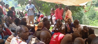 The image captures an outdoor community gathering in Kundu 1, Democratic Republic of the Congo. Approximately 30 to 40 individuals, predominantly adults of dark skin tone, are present. A man in a light blue patterned shirt and light trousers stands in the center-left, actively speaking and gesturing with both hands, addressing the assembled group. Other men are standing in close proximity, including one in a red shirt and another in an orange shirt. The majority of the attendees are seated on the ground or on low surfaces, oriented towards the standing speaker. Several individuals have their hands on their faces or heads, suggesting various states of attentiveness or emotional response. Attire is casual, consisting of assorted shirts (including one red 'Liverpool' branded shirt), trousers, and brightly patterned dresses for women. A temporary canopy, constructed from woven natural material, provides shade over the gathering. The background features lush, dense green vegetation, indicative of a natural or semi-rural environment, with glimpses of distant hillsides and faint structures in the upper right. The ground surface beneath the attendees appears to be exposed earth. A segment of corrugated metal is visible on the right edge, potentially part of a fence or structure.
