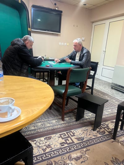 The scene depicts an indoor setting, identified as located in the city of Smederevska Palanka Municipality, Serbia. Two adult males are seated at a round table with a green felt surface, engaged in a card game.

The man on the left, with short graying hair, is wearing a black quilted jacket and dark undergarments. He is holding several cards in his hands, which are positioned over the green felt table. To his left, partially obscured by a foreground table, is a clear plastic water bottle with a blue cap.

The man on the right, also with short graying hair, wears a black leather jacket over a light-colored collared shirt. He is holding a hand of cards, his attention directed towards the game. He is seated in a dark wooden chair with a dark green upholstered back and seat, featuring decorative studs along the edges.

On the green felt table between the two men, several stacks of poker chips (red, blue, white) are visible, along with scattered playing cards, some face down and others partially exposed.

In the foreground, to the lower left, is a light-colored, round wooden table. On this table, a white ceramic cup with an orange rim and a design is resting on a matching saucer, containing a small amount of dark liquid residue.

The room features light-colored walls. A black flat-screen television is mounted on the wall above the poker table. Below the television, four light-colored square wall outlets or switches are visible. To the right of the TV, two wall-mounted coat hooks are present. A double white door with dark trim is situated on the right side of the room. Below the door, a metallic, finned radiator or heater unit is on the floor.

The floor is covered with a patterned rug featuring a central floral or medallion design in beige, brown, and blue hues, surrounded by a geometric border. Two small, dark rectangular stools are placed on the rug in the lower right portion of the image. The room is illuminated by overhead artificial lighting, with ceiling fixtures visible.