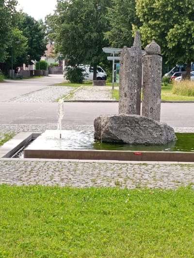 This is a full, medium shot of a public fountain with a decorative stone sculpture in the city of Brannenburg, Germany. The fountain is rectangular and filled with green water, with a thin stream of water spraying upwards from the center. A large, gray, textured rock sits in the middle of the fountain, partially submerged. Behind the fountain, two large, gray concrete pillars, textured with embedded stones, rise from the ground. The left pillar is taller and tapers to a point. The right pillar is shorter and topped with a rounded, spherical stone. The ground surrounding the fountain is paved with light gray cobblestones. Beyond the cobblestones, a paved road is visible, and further in the background, a lush green lawn with scattered white wildflowers is present. Mature trees with dense green foliage line the sides of the scene, casting shadows. In the background, a white van is parked on the road, and a light-colored bus shelter with a roof can be seen. A fence made of vertical wooden planks is also visible. The overall impression is one of a tranquil public space on a bright, overcast day.