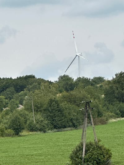 The image depicts a rural landscape with a prominent wind turbine visible in the distance, situated behind a dense line of green trees. In the foreground, a vibrant green field stretches out, bordered by a forested area. A wooden utility pole with insulators stands in the middle ground, adding to the rural feel of the scene. The sky is overcast with clouds. There are no people or animals visible in the image. The location appears to be Lysohirka, Ukraine.