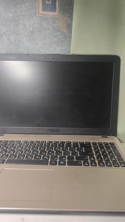 The image shows a close-up, overhead view of an open laptop computer, likely an ASUS brand, resting on a textured surface. The laptop is gold-colored with a black keyboard and screen. The screen is dark and shows some dust. The keyboard layout appears to be Cyrillic. The background consists of a patterned wallpaper and a muted green wall. The scene is indoors and appears to be a domestic or office setting. There are no people or actions depicted in the image. The location of Ukhta, Russia, is not visually identifiable from the image content.