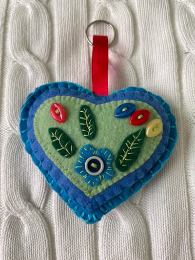 A felt heart-shaped keychain lies on a white cable-knit sweater. The keychain has layers of blue and turquoise felt, with a green felt top layer. The green layer is decorated with three oval buttons in yellow, red, and blue, three green felt leaves with yellow embroidered veins, and a blue felt flower with a blue button in the center. A silver keyring is attached to the heart by a short length of red ribbon.