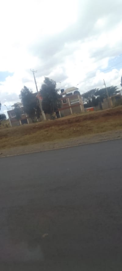 This is an outdoor, daytime photograph taken in Hells Gate ward, Kenya, under a partially cloudy sky. The lower half of the image is dominated by a dark grey paved road. Above the road, a light brown dirt verge rises slightly. In the midground, a row of multi-story buildings is visible, set amongst numerous green trees. The buildings are primarily brick-colored with lighter-colored sections and some feature flat roofs with black water tanks. A prominent utility pole with overhead lines stands among the trees. The background consists of more trees and residential-style structures under a bright, overcast sky with visible clouds. No individuals or active subjects are discernible in the scene.
