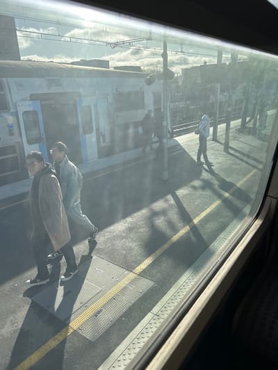The image displays a bright, sunlit railway platform scene at a station in Maisons-Alfort, France, viewed from inside a train. A light-colored RER or Transilien train, featuring "Île-de-France" branding and blue accents, is stationary on the adjacent track with one door open. The concrete platform is marked with a yellow safety line and tactile paving, and utility covers are visible. Overhead electrical lines and distant urban structures are present under a partly cloudy sky.
In the foreground, two individuals are walking from left to right across the platform. The person closer to the camera wears glasses, a dark hoodie, a light brown long coat, black pants, and black and white low-top sneakers. The second individual wears a light blue denim-like jacket and matching pants with black and white sneakers.
Further down the platform, near the open train door, several indistinct figures are observed, some appearing to be in motion. To the right, a lone individual in a light-colored hooded jacket and dark pants stands facing away from the camera, casting a long shadow. The train window exhibits reflections and glare from the intense sunlight.
