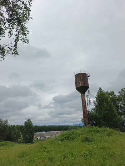 A wide shot captures a rusty water tower standing on a grassy hill, with a cluster of buildings and a dense forest in the background. The sky is overcast with heavy grey clouds. The water tower is the central subject, made of metal and showing signs of rust. A ladder is attached to its side, leading to the top. In the foreground, lush green grass covers the hill, with some bushes and trees scattered around. The middle ground features a collection of white buildings with grey roofs, possibly farm structures, nestled amongst trees. A dense line of dark green trees forms the backdrop, extending towards the horizon where distant hills are faintly visible. The overall scene suggests a rural or semi-rural setting in Medvedevka, Russia, on a cloudy day. There are no people visible in the image.