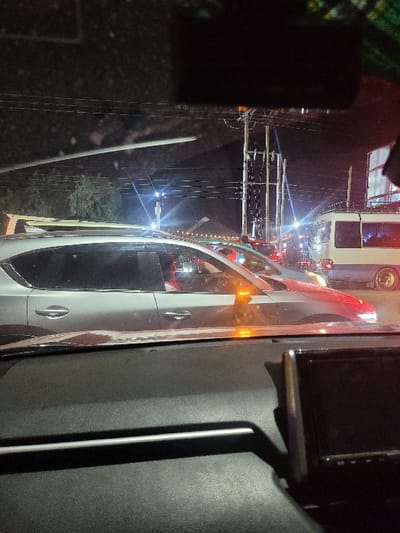 This image captures a nighttime urban street scene in Hells Gate ward, Kenya, viewed from the interior of a stationary or slow-moving vehicle. The foreground shows the dark, textured dashboard of the observer's vehicle, featuring a thin silver trim line and a black electronic display unit (possibly a navigation screen or dashcam) on the right.
Immediately beyond the windshield, a light grey SUV, likely a crossover, is positioned in the adjacent lane or lane ahead, its passenger side and front visible. Red light reflections or an interior light source are discernible within its cabin. Further to the right and slightly behind, a white and blue public service vehicle (matatu), resembling a Nissan Caravan with a roof rack, is visible. Multiple vehicles display illuminated taillights and signal lights, indicating traffic conditions.
The background is characterized by numerous utility poles with complex arrays of overhead power lines, illuminated by various artificial light sources. Bright white and yellow streetlights, vehicle headlights, and ambient illumination from distant structures or establishments contribute to the nighttime glow. Sparse dark foliage is visible on the far left. No explicit individuals or their specific actions are discernible, but the scene indicates vehicular traffic activity.