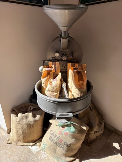 The image shows a coffee roaster filled with bags of coffee beans, and burlap sacks of beans stacked next to it. The roaster is metallic grey and has a funnel-shaped component extending from one side. The bags within the roaster are brown paper, bearing the name "Petri". Sacks of burlap next to it read "CAFES DO BRASIL" and "U/GACO Product". The setting appears to be in Tarquinia, Italy.