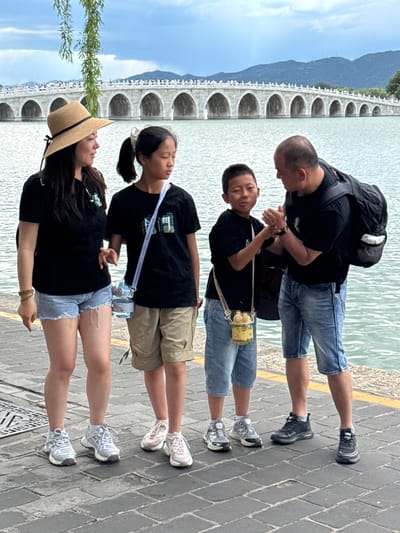 A group of four individuals, consisting of two adults (one female, one male) and two children (one girl, one boy), stands on a grey stone paved walkway adjacent to a large body of water. The background features the 17-Arch Bridge spanning across Kunming Lake in the Summer Palace, Haidian District, Beijing, China, with green mountains and a cloudy sky beyond. A tree branch with green leaves hangs into the frame from the upper left.
The female adult, positioned on the far left, wears a straw sun hat with a black band, a black short-sleeved t-shirt, light blue denim cutoff shorts, and light-colored athletic shoes. A brown beaded bracelet is visible on her left wrist. She looks towards the female child.
The female child, to the right of the female adult, wears a black short-sleeved t-shirt, khaki shorts, and light pink and white athletic shoes. Her dark hair is tied in a ponytail with a white scrunchie. A clear cylindrical water bottle hangs from a white strap around her neck. She looks downwards and slightly to the right.
The male child, to the right of the female child, wears a black short-sleeved t-shirt, light blue denim shorts, and grey and white athletic shoes with yellow accents. A yellow animal-shaped water bottle hangs from a strap around his neck. He is facing the male adult, with his hands clasped together near the male adult's hands.
The male adult, on the far right, has a bald head and wears a black short-sleeved t-shirt, blue denim shorts, and dark athletic shoes. A black backpack is worn on his shoulders. He leans slightly towards the male child, engaging in a hand interaction with the child, his hands also clasped together.
The walkway includes a metal grate on the left and a yellow painted line along its edge on the right. The water surface exhibits minor ripples.