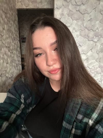 A young woman with long, dark brown hair is shown in a close-up, selfie-style photograph. She is wearing a black t-shirt and an open, dark green and black plaid flannel shirt. Her face is partially turned to the side, with her right eye and lips clearly visible. Her left eye is looking directly at the camera. The background consists of wallpaper with a repeating floral pattern. The image appears to have been taken indoors. The location in the city of Osentsy, Russia, is not identifiable from the visual information provided.