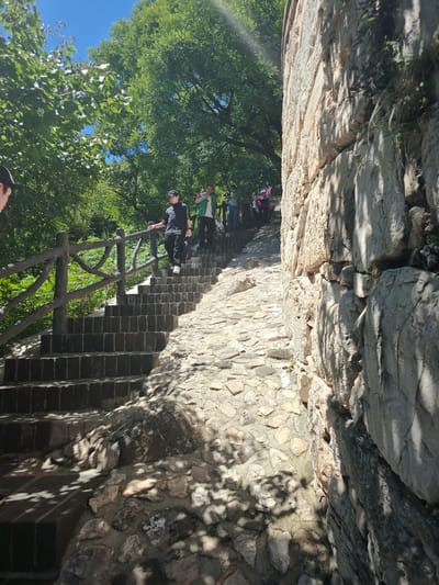 The image depicts a stone staircase leading uphill, bordered by a rustic wooden railing on the left and a rough-hewn stone wall on the right. Sunlight filters through lush green trees, casting dappled shadows on the stone steps and pathway. Several individuals are visible ascending the stairs. A person in a black uniform and cap walks down the steps, holding onto the railing. Further up, other people, some wearing casual clothing and bright colors, are also walking. The overall scene suggests an outdoor location with historical or natural significance, possibly a tourist attraction. Based on the context of similar images and the architectural style, this location is likely within the Mutianyu section of the Great Wall in Huairou District, China, known for its well-preserved and scenic portions.