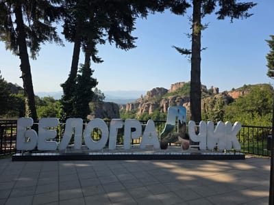 The image shows a large, white, three-dimensional inscription spelling out "BELOGRADCHIK" in Cyrillic letters against a backdrop of the Belogradchik Rocks in Belogradchik, Bulgaria. The inscription is positioned on a paved area, with trees and a metal railing in the foreground. The Belogradchik Rocks, a series of unusual rock formations, are visible in the background under a clear blue sky. One of the letters in the inscription is decorated with an image of hot air balloons and part of the rock formations. The overall scene appears to be a viewpoint or tourist attraction.