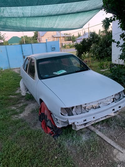 A white car body is the central object in this outdoor scene. The car is unfinished, with its hood open and front end disassembled, revealing exposed engine components and wiring. It appears to be undergoing repairs or restoration. A red and black tarp or cover is partially visible beneath the front of the car. The car is situated on a patch of ground with dry soil and patches of green grass.
To the left of the car is a tall, green mesh tarp or shade cloth, providing cover. In the background, there are hints of a rural or semi-rural setting. Buildings with light-colored walls and a blue structure are visible, suggesting a residential or industrial area. Trees and foliage are also present. The lighting suggests it is either early morning or late evening, with a soft, diffused light. The sky appears hazy or overcast.
The overall impression is one of a project in progress, outdoors, in what seems to be a location in Зертас ауылдық округі, Kazakhstan. There is no visible text in the image.