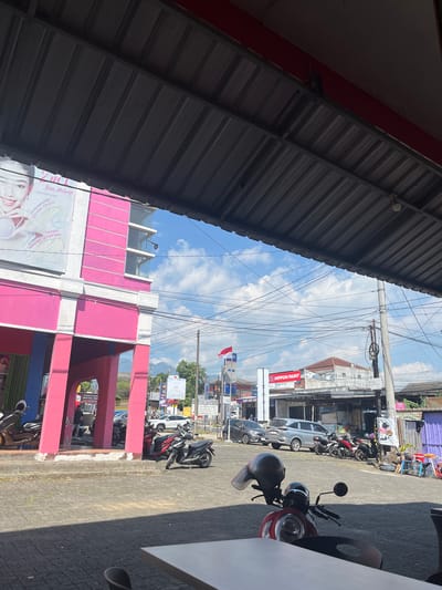 The image depicts a street scene in Tampingan, Indonesia, on a sunny day. The foreground is dominated by the underside of a corrugated metal roof, casting a shadow over part of the frame. To the right, a red motorcycle with a black helmet on its seat is parked on a paved area. Beyond the motorcycle, a row of buildings lines the street, featuring a prominent pink structure. Various motorcycles and a few cars are parked along the roadside.

Several signs are visible, including one for "NIPPON PAINT" and another advertising "Base Makeup" with claims of high coverage and all-day oil control. The sky is blue with scattered clouds, suggesting fair weather. Power lines crisscross the sky above the street. The overall impression is of a typical commercial area in a Southeast Asian town during daylight hours. The presence of parked vehicles and shops suggests ongoing commercial activity.