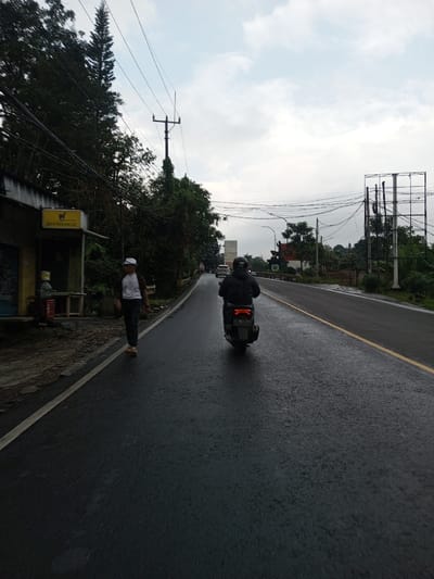 The scene depicts an overcast day on a wet asphalt road in Dawathang_Dorjibi_Kashingtsawa, Bhutan. In the foreground, a dark-colored scooter, featuring a red taillight, is traveling away from the camera on the right side of the road. The operator wears dark clothing and a dark helmet with a white "007" logo on the back. To the left, a male pedestrian walks in the same direction, wearing a white t-shirt with brown accents, dark trousers, light-colored shoes, and a white baseball cap worn backward.

The left side of the road features a raised dirt/concrete shoulder alongside a single-story building with a blue roll-down shutter and a yellow sign above displaying the word "ROONUNCIA" and a cup-like symbol. A small wooden table or bench is visible outside this structure. Dense green foliage, including a tall coniferous tree, lines this side of the road. Overhead, multiple utility poles with a complex network of black electrical or communication wires traverse the sky, which is heavily clouded.

The road itself is dark and visibly wet, suggesting recent precipitation, and is delineated by a white solid line on the left and a faded yellow line on the right. In the distance on the right, a large, empty metal billboard frame stands prominently. Further down the road, additional white vehicles are discernible, along with a structure featuring a red roof and a dark monument or statue in front of it.