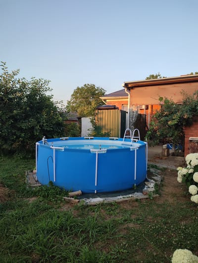 This media file shows an above-ground, round, blue swimming pool filled with clear water. The pool has white support poles and a white ladder. It is set up in a backyard with green grass and trees. In the background, there is a brick house with a brown roof, a green fence, and other vegetation. The scene appears to be a residential backyard in Abinsk, Russia. The lighting suggests it is late afternoon or early evening, with the sun casting a warm glow on the house. The weather seems pleasant, suitable for outdoor activities. There are no people visible in the image. The overall mood is tranquil and suggests a peaceful, domestic setting. No visible text is present in the image.