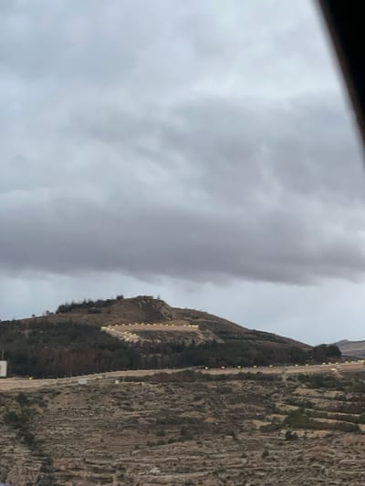 The image captures a rugged, expansive hillside landscape within Dummar Municipality, Syria, under an overcast sky filled with dense grey clouds. The primary subject is a large hill that occupies the central and upper portions of the frame. On the upper and mid-slopes of this hill, a prominent series of bright, yellow-white artificial lights are arranged in multiple linear segments, forming a distinct, illuminated path or structure that meanders across the terrain. These lights are evenly spaced and project illumination onto the immediate ground. Patches of dark green trees and shrubs are visible on the hillside, interspersed with areas of sparse, brownish vegetation and exposed earth. The lower sections of the hill feature discernible terraced land formations. To the far left of the frame, a white, rectangular structure with a flat roof is partially visible at a lower elevation on the hillside. Another horizontal line of similar yellow-white lights is visible along a lower terrace or road in the midground. No individuals or specific human activities are observable within the scene.