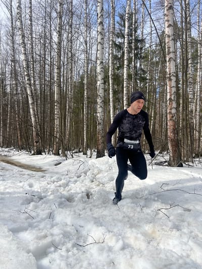 A man is positioned in a winter environment, possibly during a race or training session, based on his attire and race number "73." He is wearing black athletic clothing consisting of a beanie, long-sleeved shirt, pants, and gloves. The man is standing on a snowy terrain. Behind him is a dense forest of birch trees, with their characteristic white bark and thin, bare branches reaching towards the sky. The sky is partially visible through the branches, appearing to be a light blue color. The presence of birch trees suggests this is in Russia, but there is no definitive evidence of the city of Zelenodolsk, Russia in the image.