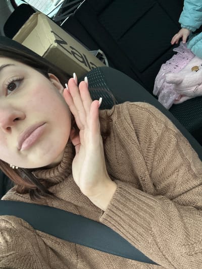 The scene is set inside a vehicle, in Reichstett, France. A young adult female, positioned in the foreground and wearing a brown knitted turtleneck sweater, is secured by a black seatbelt across her chest. Her left hand is raised, with fingers slightly spread and nails featuring white tips, partially obscuring the lower-left side of her face. Her head is tilted slightly to the right, revealing part of her face, including her left eye, nose, and slightly parted lips. Her hair is dark brown. In the background, behind her, a brown cardboard box with the visible text "Zala" is present on a dark seat. To the right, partially visible on an adjacent dark seat, is the arm and hand of a second individual, likely a child, wearing a light blue puffer jacket. Adjacent to the child's arm is a light pink, plush bag, possibly a backpack, with a unicorn design and a sheer, sparkly overlay. The vehicle interior features black fabric upholstery.