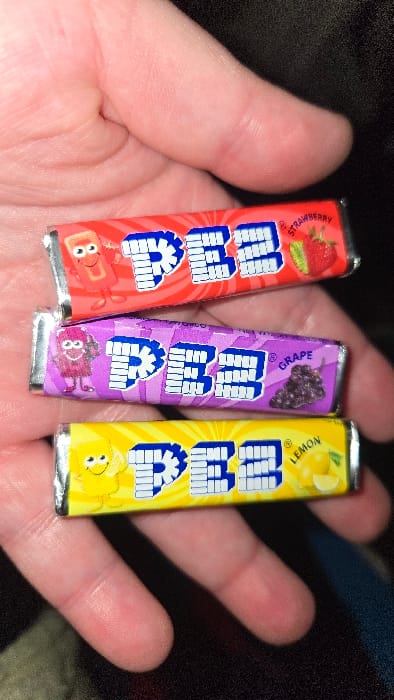 A hand holds three wrapped PEZ candy packages. The flavors are strawberry (red), grape (purple), and lemon (yellow). Each wrapper has the PEZ logo in blue and white. A small cartoon character related to the flavor is printed at the bottom of each package. The packages are held upright and close together. The person's skin tone is pale. A dark background is slightly visible behind the hand.