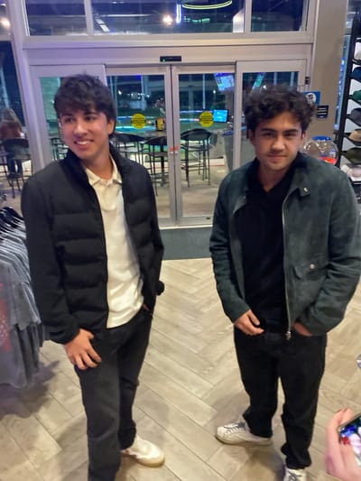Two young men stand inside a building with a glass facade. One wears a gray-green jacket and black shirt and pants, while the other is in a black puffer jacket over a white shirt, also with black pants. Both are wearing white sneakers. The floor has a light brown herringbone pattern. Through the glass, tables and chairs are visible, along with outdoor lights and a display screen. A rack of clothing is visible at the bottom of the frame. Someone is holding a phone in the upper right corner.