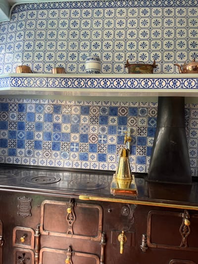 The image shows a kitchen stove with a brass faucet in the kitchen of Claude Monet's home at 99 Rue Claude Monet, Giverny, France (latitude: 49.075, longitude: 1.534). The stove is dark brown and made of cast iron. The stovetop is black and has two round burners. The stove has a brass trim and several brass knobs and handles. The stove is set against a wall that is tiled in blue and white ceramic tiles. The tiles are arranged in a pattern of squares and rectangles. The wall above the stove is tiled in a similar pattern. The tiles feature blue and white geometric designs. There are three objects on the shelf above the tiles: a white ceramic jar, a small brass container with a hinged lid, and a copper pot. The wall above the stove has a pale green trim and the ceiling is white.  The image is taken from a slightly elevated angle, looking down at the stove and the wall behind it. The lighting is soft and natural. The image is in focus and well-composed.