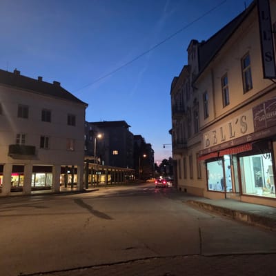 The image depicts a street scene in Piešťany, Slovakia, during twilight. The sky is a deep blue, with faint wisps of clouds and what appear to be contrails from an aircraft. The street is paved and shows signs of wear, with visible cracks.

On the right side of the street, a building with an ornate facade features a storefront labeled "BELL'S" in large, light-colored letters. Above and to the right of "BELL'S" is a vertical sign with the letters "L" and "S". The storefront has large glass windows displaying mannequins dressed in clothing. Awning fabric is visible above the windows, featuring a striped pattern. Below the main "BELL'S" sign, smaller text indicates "Laura Biagiotti", "VALENTINO", and other brand names.

On the left side of the street, a more modern building with a lighter facade is visible. It has a covered walkway with supporting columns, beneath which are shopfronts with illuminated interiors. One of these shopfronts displays items through its windows, and another features a sign for "CASINO".

In the distance, several multi-story buildings are visible, some with illuminated windows. Streetlights are present along the road. A few cars are on the street; one car, a silver hatchback, is in the foreground with its brake lights illuminated. Another car, a yellow sedan, is further down the street. The scene suggests a commercial area of the city as evening approaches.