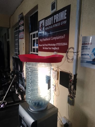 An interior view of the Body Prime Health & Fitness Gym in Njoro ward, Kenya, is depicted. A grey water dispenser, topped with a large, clear plastic water bottle containing water, is centrally positioned. A red, elongated tray-like object rests horizontally on the inverted water bottle.
On the wall behind the dispenser, a prominent black and red banner sign for "BODY PRIME HEALTH & FITNESS GYM" is displayed, featuring contact details for feedback. To its immediate left, a smaller sign lists pricing: "Session Ksh 100" and "Monthly Ksh 2,500". Further left, white-framed window-like structures are visible, and in the dark background, indistinct gym equipment, including what appears to be a purple kettlebell, is present.
To the right of the main gym sign, a white pillar supports multiple electrical wires, cables, and various mounted electronic components, including power adapters and what resembles a network device. A power outlet is visible near the bottom right. A white and blue sign, partially visible on the far right, advertises "DRINKING WATER SERVICES". Artificial illumination causes a bright reflection on the water bottle and lights the immediate area. No individuals are visible in the scene.