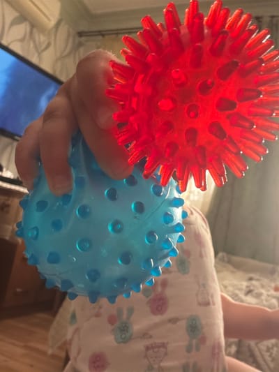 The image shows a close-up of a child's hand holding two spiky sensory balls, one red and one blue. The red ball is partially visible in the upper right, while the blue ball is in the foreground, held by the child's fingers. The child is wearing a light-colored shirt with a pattern of cartoon rabbits. In the background, a television screen is visible on the left, and a bed with patterned bedding is on the right. The scene appears to be indoors, possibly in a child's bedroom or living area. The lighting is somewhat dim, with light reflecting off the plastic balls. The image is taken from a low angle, looking up towards the child and the toys. It is not possible to determine the specific location within Komsomolskiy, Russia based on the visual information provided in the image.