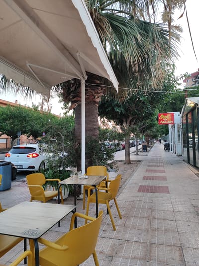 An outdoor seating area of a restaurant or cafe is depicted in El Campello, Spain, likely in the late afternoon or early evening as indicated by the soft, warm light. The scene is a sidewalk lined with tables and bright yellow chairs. The tables are made of wood with metal frames, and one of them displays a Heineken "Pura Malta" logo. On one of the tables, there are glasses and a small plate with what appears to be olives.
Behind the seating area, a sidewalk paved with square tiles and accented with rectangular red tiles leads into the distance. Palm trees and other lush green foliage line the sidewalk, providing shade and a tropical ambiance. A white car is parked on the street to the left, with its brake lights illuminated. Buildings, typical of a Mediterranean town, are visible in the background. A red and yellow sign that reads "Rico Kebab-pizzeria" is prominently displayed on the facade of a building along the sidewalk. A few people are seen walking in the distance, suggesting a relaxed evening atmosphere. The overall mood of the image is peaceful and inviting, typical of a pleasant evening in a Spanish coastal town.