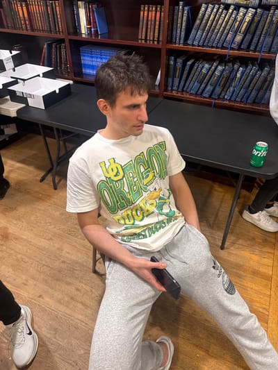 The image captures an indoor scene in Bergenfield, United States, appearing to be a library or study hall. A young adult male is seated on a chair in the lower-center, angled towards the right side of the frame. He wears a white t-shirt with a green and yellow "U OF OREGON DUCKS" graphic and light grey Adidas sweatpants. His right hand holds a dark smartphone, resting on his right thigh, while his left hand is in his left pocket. Behind him, a black rectangular folding table extends across the frame. On the table's left, several stacked white cardboard boxes with black edges are visible, one displaying the inverted text "A TASTE OF HOME." On the table's right, a green Sprite soda can rests. The background features dark wooden bookshelves completely filled with books, many uniformly bound in blue with gold-colored spine text. The floor is composed of light brown wooden planks. Partial views of two other individuals are present: a person wearing white Nike sneakers on the far left, and another person wearing white Nike Air Force 1 sneakers on the far right.
