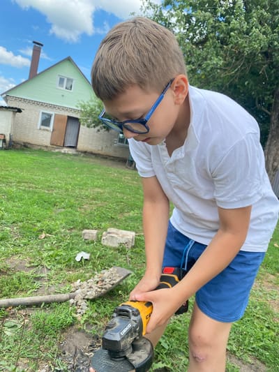 In this vertical outdoor shot, a young boy is seen from the chest up, wearing a white t-shirt and blue shorts. He is looking down and focusing intently on an angle grinder in his hands. The grinder is yellow and black, with the brand name "DEKO" visible on it. The boy's hair is short and light brown, and he is wearing blue-rimmed glasses. His right knee, visible in the frame, has a bandage or scab.
The scene appears to be a yard with green grass and some scattered debris, including pieces of concrete. In the background, a light green house with a brown-shingled roof is visible, along with a brick house and a chimney. The sky is blue with white clouds. The overall impression is a sunny day, possibly late afternoon or early evening, given the angle of the light. The boy's focused expression suggests he is engaged in a task, possibly construction or repair work, as indicated by the angle grinder and debris. The location context provided is Лебяжье, Russia.