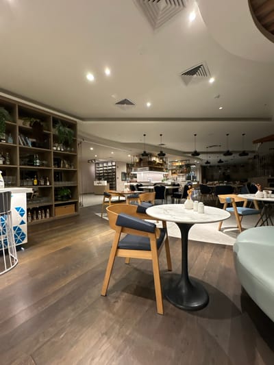 The image depicts the interior of a contemporary dining or lounge area within a commercial establishment located in Zeytinburnu, Turkey. The foreground features dark wood plank flooring, which transitions to a lighter, possibly tiled or carpeted area under a central seating arrangement. The ceiling is white, equipped with multiple recessed spotlights and several square air vents, from which dark-colored pendant lights are suspended.

On the left side of the foreground, a tall, multi-compartment wooden shelving unit displays various bottles, glassware, and potted plants. A white-framed bar stool with a base featuring a blue and white geometric pattern is partially visible. In the central foreground, a round table with a white marble-patterned top and a dark, trumpet-shaped base holds two white cylindrical shakers and a small white vase containing dried flora. Adjacent to this table is a light wooden chair with a dark blue upholstered seat and backrest. A light blue upholstered bench or sofa is partially visible on the far right.

The midground and background reveal a larger dining area furnished with numerous tables and chairs, some matching the light wood and blue upholstery scheme. A bar or service counter with illuminated shelving is visible in the midground, featuring a sign that appears to read "magnolia" above it. Further back on the left, a dark-colored wine rack wall is present. No individuals are present in the scene.