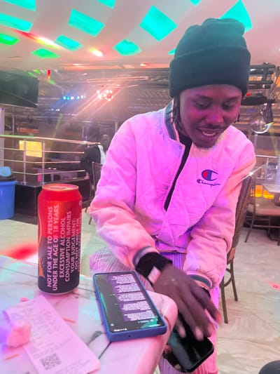 The image captures a vibrant night scene indoors, likely a bar or club, in Kimorori, Kenya. The main subject is a young man, smiling broadly, seated at a table. He wears a dark beanie and a light-colored, possibly off-white or very pale pink, quilted bomber jacket featuring a "Champion" logo on the left chest. His right hand is visible, holding a black smartphone, while his left wrist has a dark band.
On the table in front of him, illuminated by ambient lighting, are several items. A red beverage can stands prominently, displaying extensive warning text. Another smartphone, encased in a blue protective cover, lies flat on the table, its screen displaying a block of text. Also on the table are a few crumpled receipts or pieces of paper.
The background features dynamic lighting, with large, bright teal-colored square lights on the left wall and various other colored lights (pink, red, blue) illuminating structures that suggest a stage, bar, or entertainment area. A small disco ball is faintly visible in the upper background, reflecting specks of light. The overall atmosphere appears festive and social.
Visible text includes:
* On the jacket: "Champion"
* On the red can: "NOT FOR SALE TO PERSONS UNDER THE AGE OF 18 YEARS. EXCESSIVE ALCOHOL CONSUMPTION IMPAIRS YOUR JUDGEMENT: DO NOT DRIVE OR OPERATE HEAVY MACHINERY"
