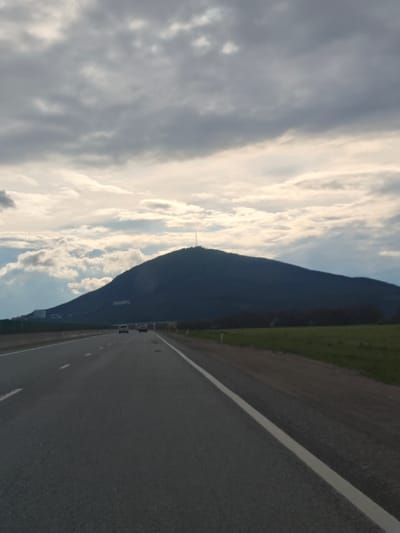 The image shows a highway leading towards a mountain under a cloudy sky. The road surface is dark gray asphalt with a white lane marking on the right. Several cars are visible in the distance. The mountain, dark green in color, has a tall tower-like structure on its peak. A green field is present on the right side of the road. There is a sparse line of trees bordering the road. The location appears to be near Konstantinovskaya, Russia, based on the landscape characteristics.