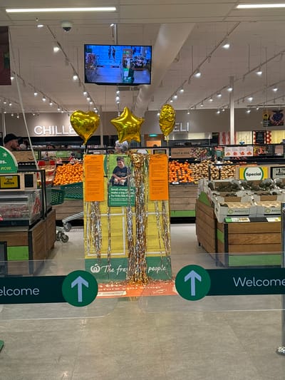 This image depicts the interior of a Woolworths supermarket in Esperance, Australia, likely near its entrance or produce section. The view is from a low angle, looking inward.

In the immediate foreground, a clear barrier with green "Welcome" signage and white upward-pointing arrows indicates an entry point. The floor is light grey tiled.

Centrally, a promotional display stands adorned with golden tinsel streamers. Three helium-filled Mylar balloons float above it: one heart-shaped and two star-shaped, all golden. The display features orange and green signage with text, prominently displaying a photograph of a young person in a wheelchair and the slogan "Small changes can make a big change for kids like Ayan." The Woolworths logo and slogan "The fresh food people" are visible at the base of this display.

To the left of the display, the "CHILLED" section sign is visible. Produce bins filled with oranges in green crates are present. A male customer with a dark cap is partially visible on the far left.

To the right of the display, the "DELI" section sign is partially visible. Further right, produce bins contain oranges, potatoes, and avocados, some labeled "AVOWEST." Green "Special" signs are visible, one indicating a price of "$1.70."

Overhead, a white suspended ceiling features numerous fluorescent light fixtures and visible air conditioning ducts. Two dome-shaped security cameras (CCTV) are mounted on the ceiling. A large flat-screen television is suspended, displaying a scene of multiple individuals engaged in a water-based activity, possibly in a swimming pool or at a waterfront. A person with grey hair is partially visible behind the central display, facing away from the camera.